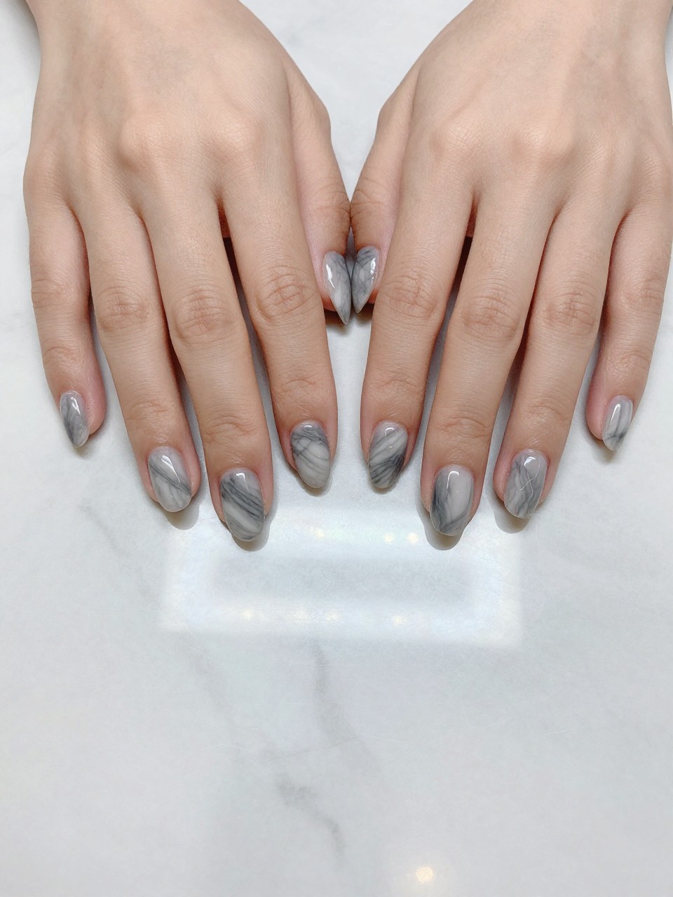 Top-down view of hands on a polished Carrara marble countertop, the nails mirroring the surface beneath. The squoval nails feature a realistic gray and white marble swirl pattern. The lighting is bright and clear, showing every delicate vein.