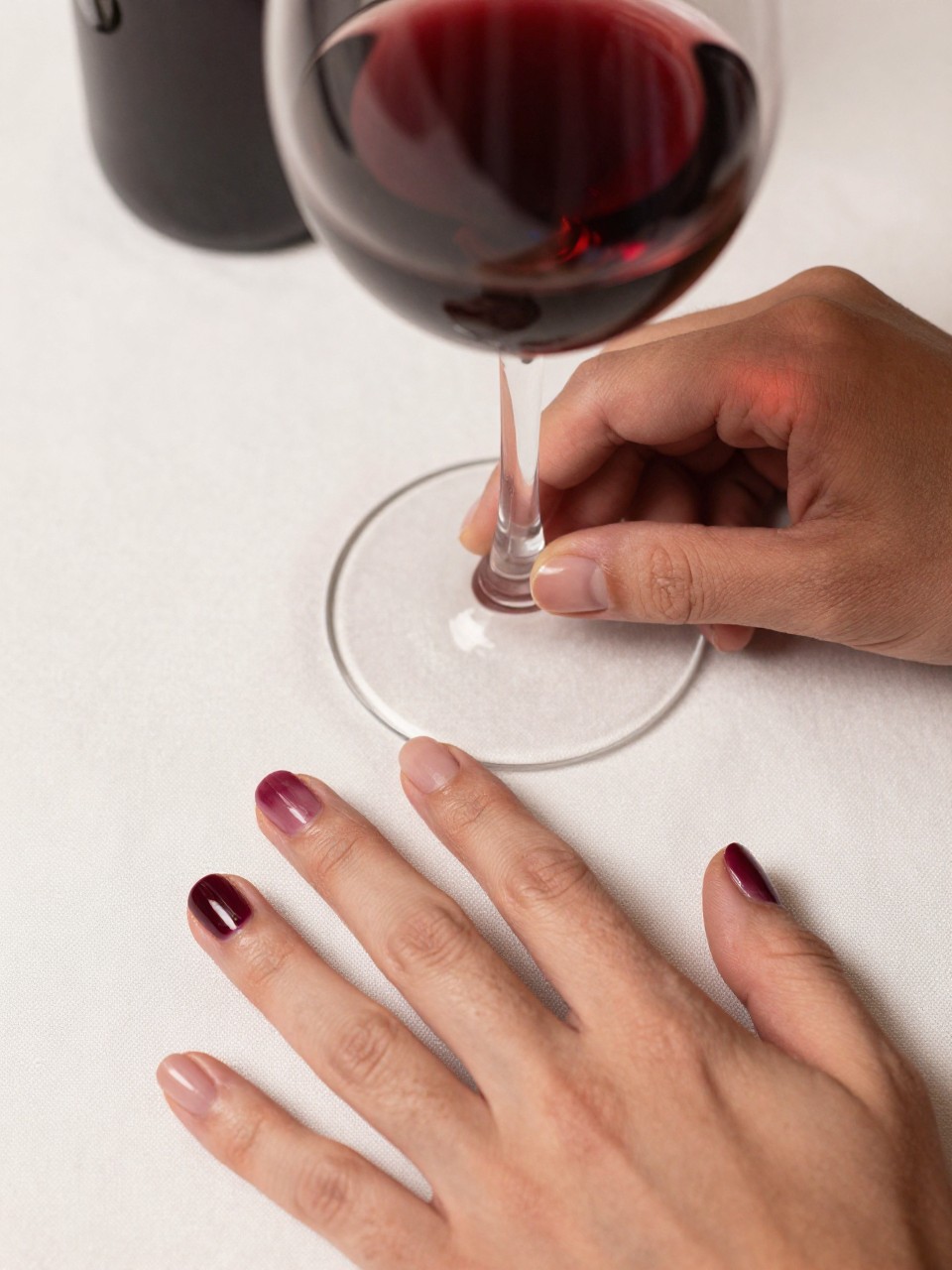 Overhead shot of hands on a white linen tablecloth next to a glass of red wine. The nails are natural and oval. A blurred, deep burgundy "stain" sits artfully on each nail bed. The lighting is warm and intimate, like a dinner party.