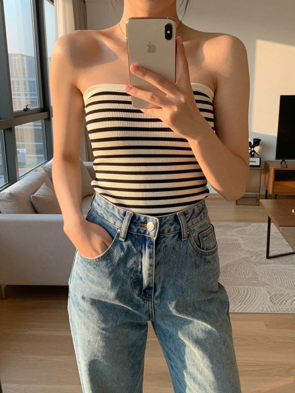 iPhone mirror selfie of a woman in a striped boatneck top tucked into high-waisted jeans, holding her phone at chest level with one hand casually in her pocket, cropped at NECK so face doesn't show, in a sunlit city apartment, evening golden hour, iPhone photo quality.