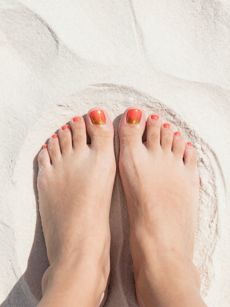  Top-down view of bare feet with perfectly pedicured toes, heels dug into warm, white sand. The toenails are a glossy coral. A gradient of gold glitter starts at the tip and fades halfway down the nail. Sunlight sparkles on the glitter.