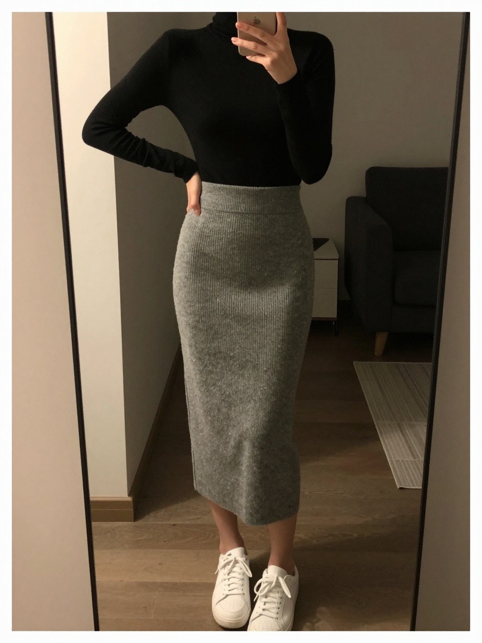 iPhone mirror selfie of a woman in a black turtleneck tucked into a gray knit midi skirt with white sneakers, standing with weight shifted to one hip, phone held low, cropped at NECK so face doesn't show, in a city apartment, warm moody lighting, iPhone photo quality.