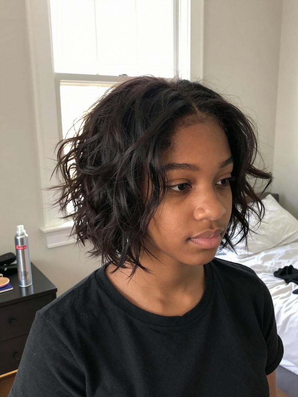 Photo of a american black teenage girl wearing a messy textured bob, angled front view showing the undone texture, setting: a bedroom with natural light from a window, lighting: bright, soft window light, containing a can of dry shampoo on a nightstand, iPhone photo quality.