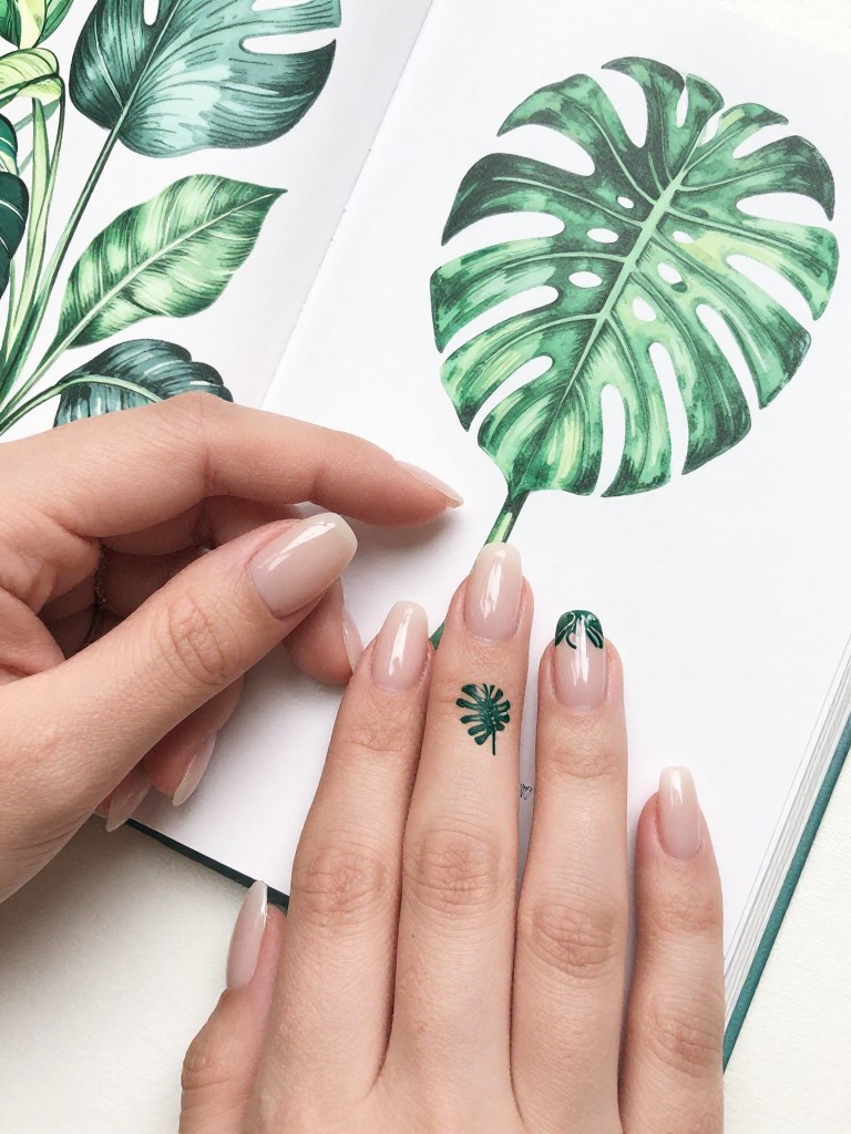 Top-down view of hands with a botanical illustration book open nearby. The nails have a sheer nude gel base. One detailed, line-art style monstera leaf is painted in dark green on each nail. The look is modern, crisp, and slightly edgy.
