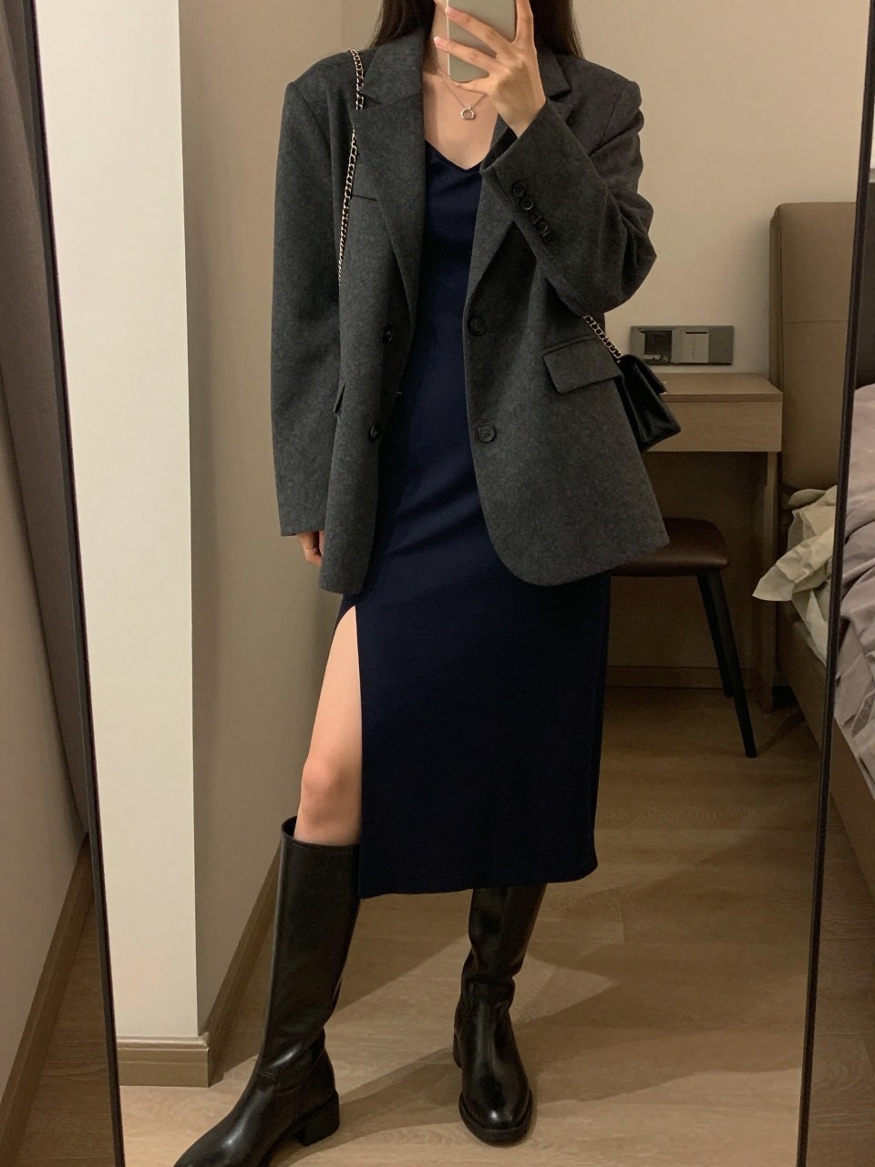 iPhone mirror selfie of a woman wearing the charcoal blazer over a navy slip skirt with knee-high boots and a chain-strap bag, posed with her weight on one leg, cropped at NECK so face doesn't show, in a city apartment, warm moody lighting, iPhone photo quality.