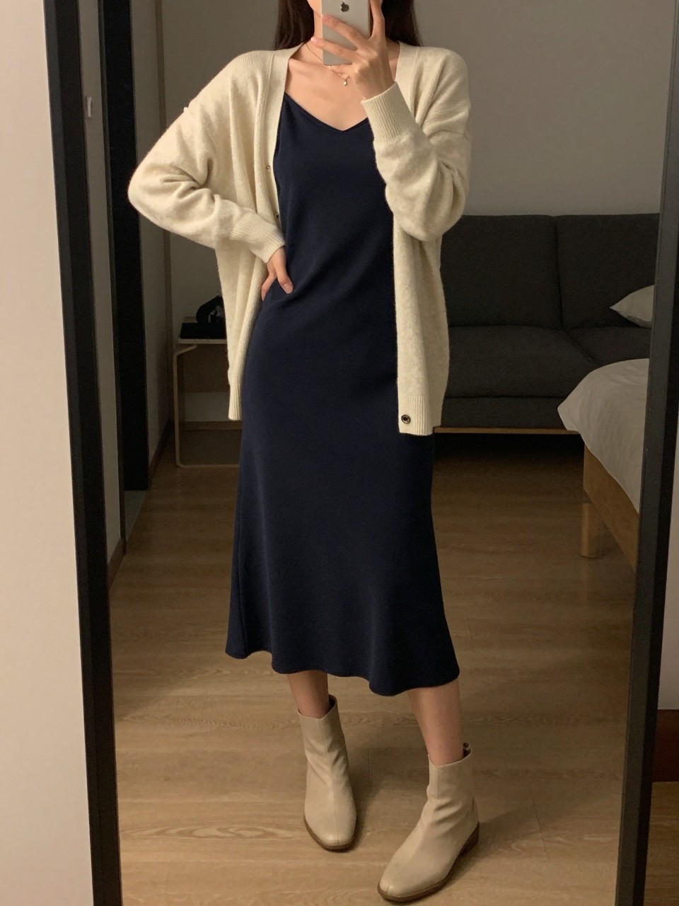 iPhone mirror selfie of a woman in a navy slip dress with a cream cardigan layered over it and flat ankle boots, standing with weight shifted to one hip, phone held low, cropped at NECK so face doesn't show, in a city apartment, warm moody lighting, iPhone photo quality.
