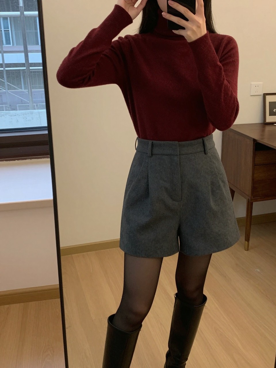 iPhone mirror selfie of a woman in a burgundy turtleneck with gray tailored shorts, black tights, and knee-high boots, adjusting a matching beret, cropped at NECK so face doesn't show, in a city apartment, warm moody lighting, iPhone photo quality.