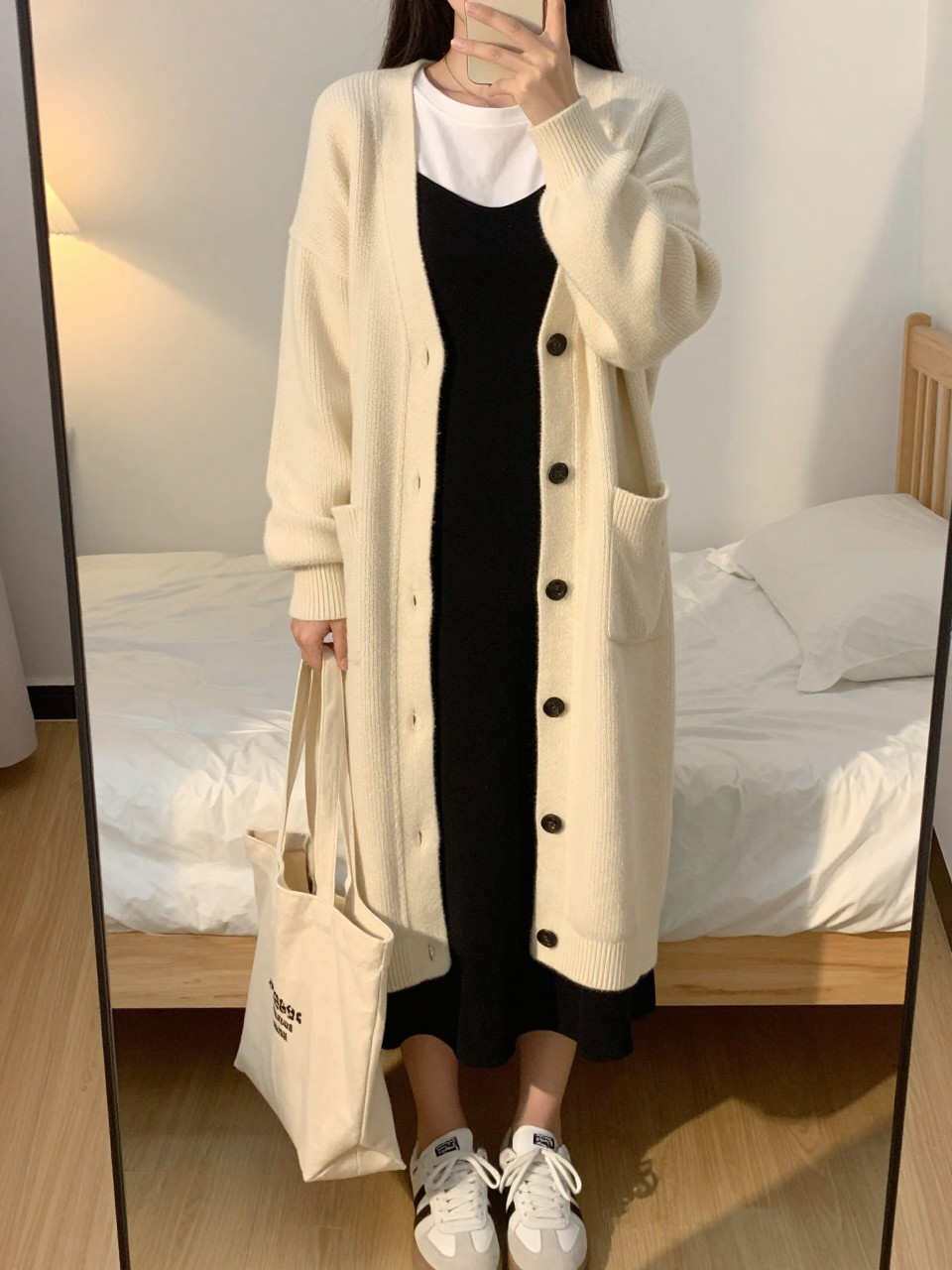 iPhone mirror selfie of a woman wearing the long cream cardigan open over a white tee and black slip skirt with white sneakers, holding a canvas tote, posed with her hand adjusting her collar, cropped at NECK so face doesn't show, in a feminine-themed bedroom, evening golden hour, iPhone photo quality.