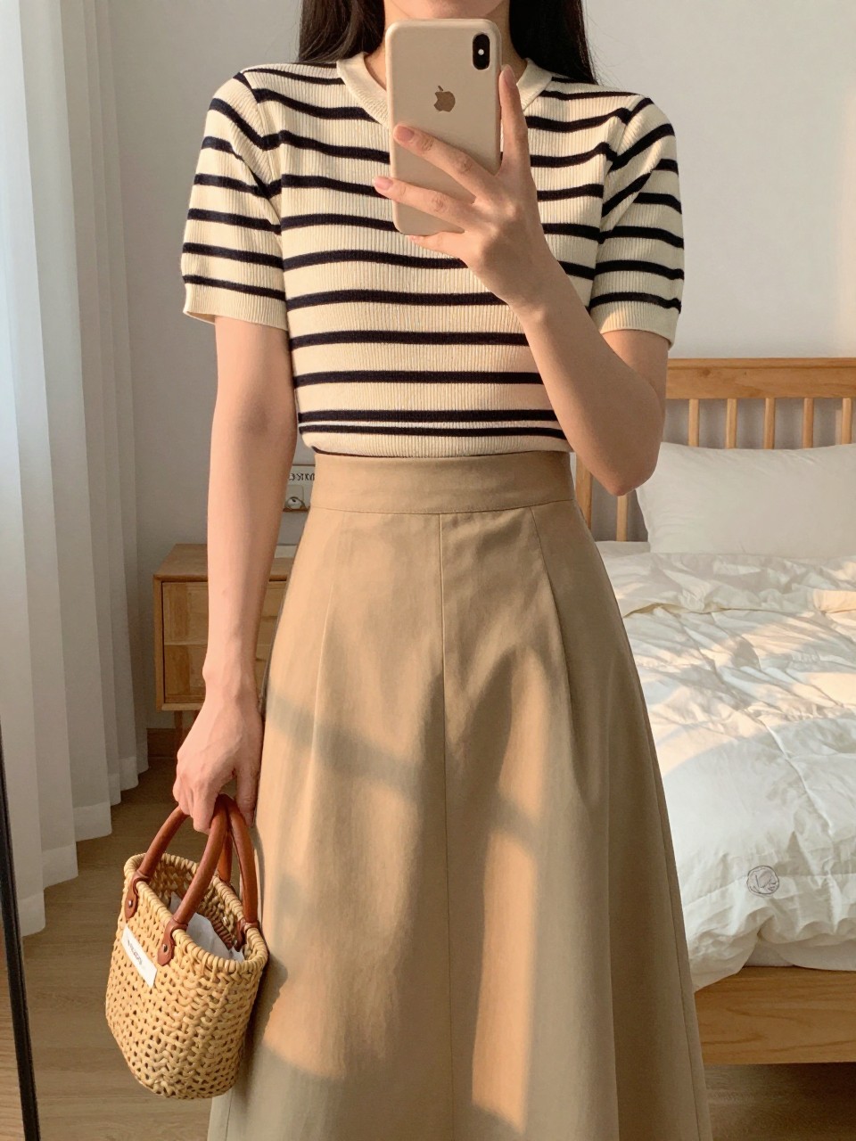 iPhone mirror selfie of a woman in a striped top tucked into an A-line skirt, holding her phone at chest level while holding a basket bag, cropped at NECK so face doesn't show, in a feminine-themed bedroom, evening golden hour, iPhone photo quality.