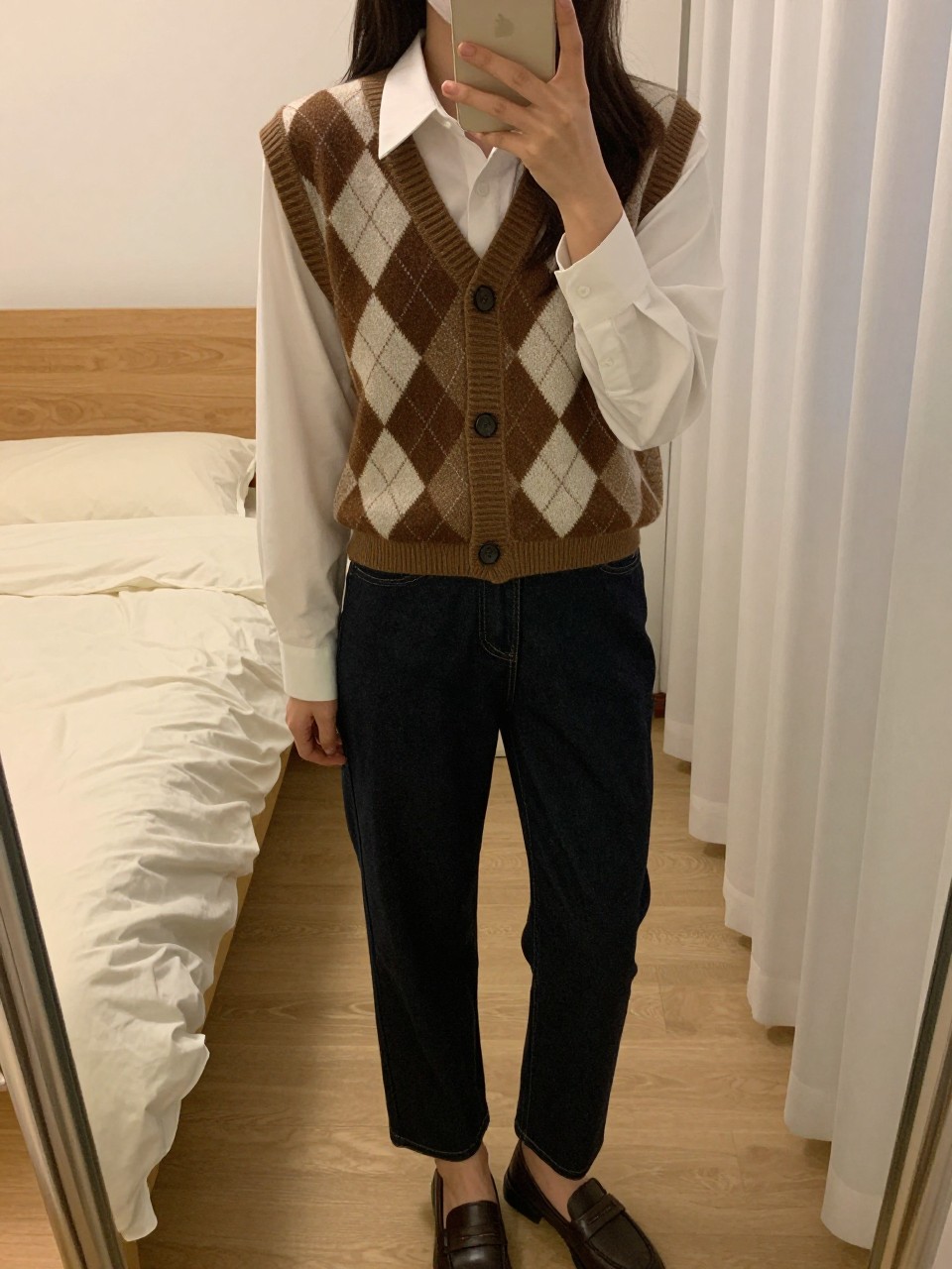 iPhone mirror selfie of a woman in an earth-tone argyle waistcoat over a white button-down with dark straight jeans and loafers, looking at her phone, cropped at NECK so face doesn't show, in a feminine-themed bedroom, warm moody lighting, iPhone photo quality.