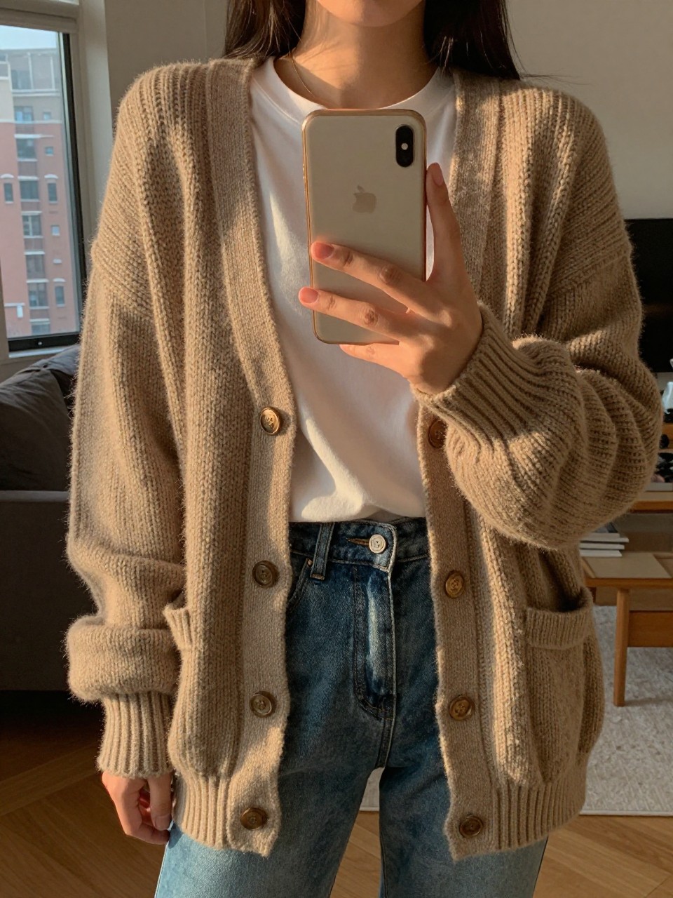 iPhone mirror selfie of a woman in a chunky cardigan over a tee and jeans, holding her phone low, one hand holding the edge of the cardigan, cropped at NECK so face doesn't show, in a sunlit city apartment, warm moody lighting, iPhone photo quality.