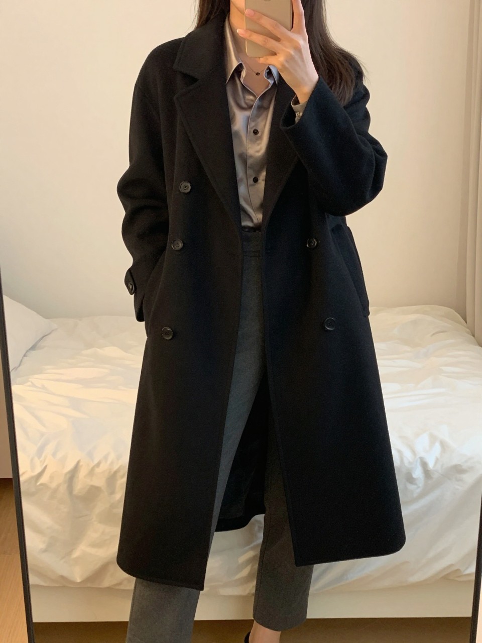 iPhone mirror selfie of a woman wearing a tailored black coat over a silk blouse and trousers, standing with her weight on one leg and phone at her side, cropped at NECK so face doesn't show, in a bright feminine-themed bedroom, evening golden hour light, iPhone photo quality.