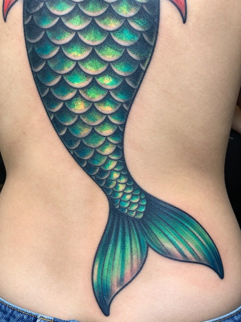 "Detail focus on the mermaid's tail tatoo transitioning into scales near the lower back. The scales have an iridescent color shift (green/blue), captured under light that makes them shimmer, with wave patterns intertwining. This must be a natural and realistic tattoo design
