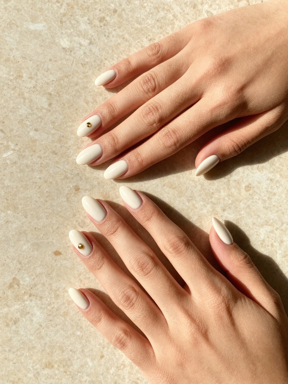 Overhead, top-down view of feminine hands with almond-shaped nails resting on a sun-warmed, travertine marble surface. The nails are a soft, creamy white. A single, burnished gold dot sits near the cuticle of each ring finger. The lighting is bright and warm, casting soft shadows.