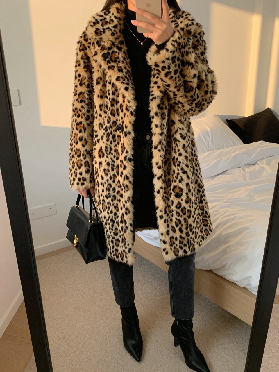 iPhone mirror selfie of a woman wearing the leopard faux fur coat over a black sweater and dark jeans with pointed-toe ankle boots, holding a mini black bag, posed with her hand in her coat pocket, cropped at NECK so face doesn't show, in a feminine-themed bedroom, evening golden hour, iPhone photo quality.