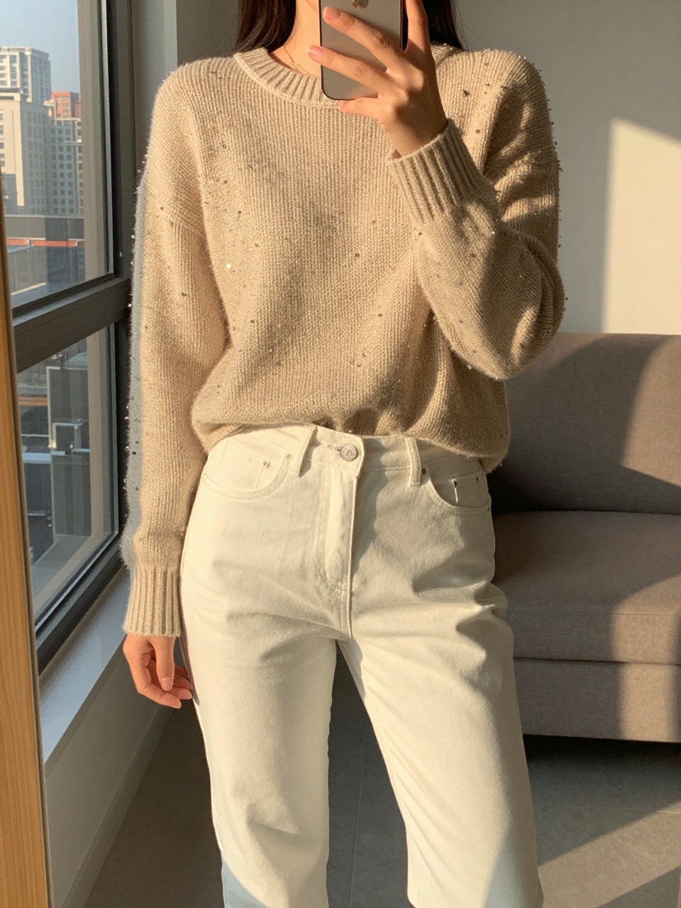 iPhone mirror selfie of a woman in a beaded sweater and white jeans, holding a cup, cropped at NECK so face doesn't show, in a city apartment, golden hour sunlight, iPhone photo quality.