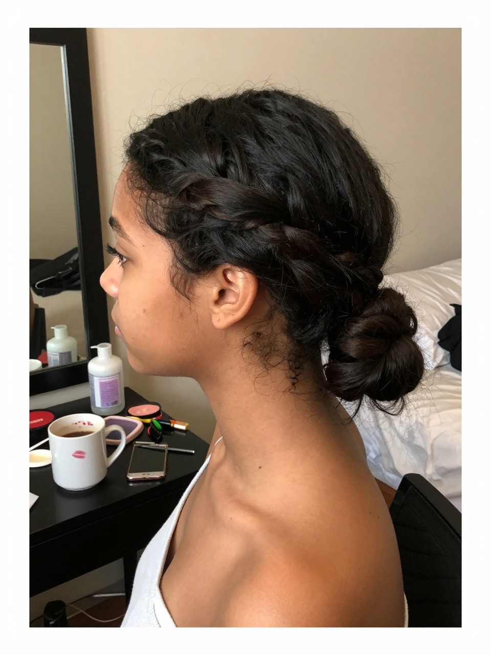 Photo of a biracial teen girl wearing a twisted low ponytail, side profile view showing the twisted detail, setting: a bedroom with a mirror and getting-ready clutter, lighting: soft, warm indoor lighting, containing a coffee mug with a lipstick stain on the rim, iPhone photo quality.