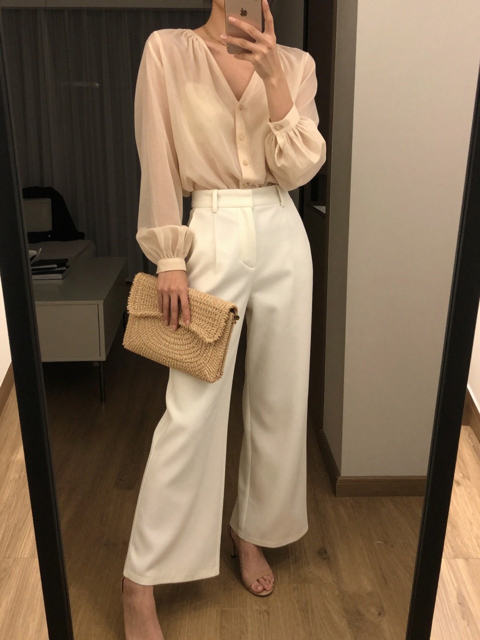 iPhone mirror selfie of a woman in an ivory sheer blouse and white wide leg pants with nude heeled sandals, holding a raffia clutch, slight pose with phone, cropped at NECK so face doesn't show, in a dim city apartment, warm moody lighting, iPhone photo quality.