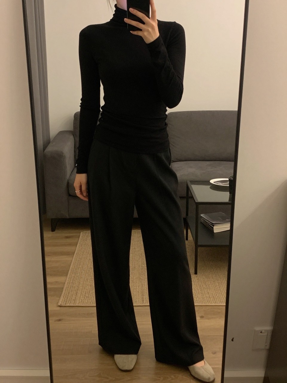 iPhone mirror selfie of a woman in a fitted black turtleneck tucked into wide leg pants, standing with phone at waist level, one foot slightly forward, cropped at NECK so face doesn't show, in a cozy city apartment, warm moody lighting, iPhone photo quality.