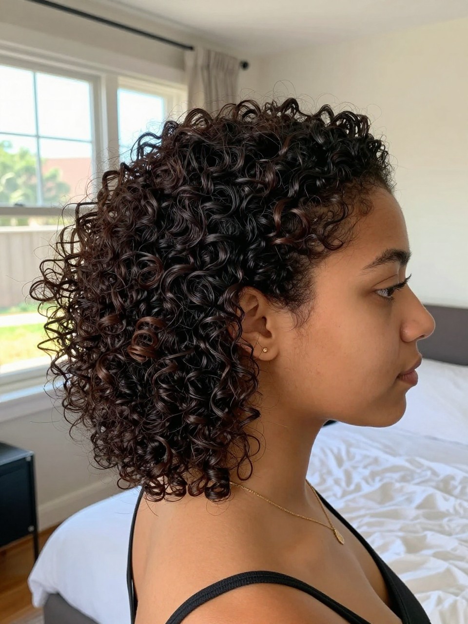 Photo of a woman wearing a curly low pony with volume, side profile view showing lifted crown and sleek front, setting is a sun-drenched bedroom with natural window light, containing a simple gold necklace visible, iPhone photo quality.