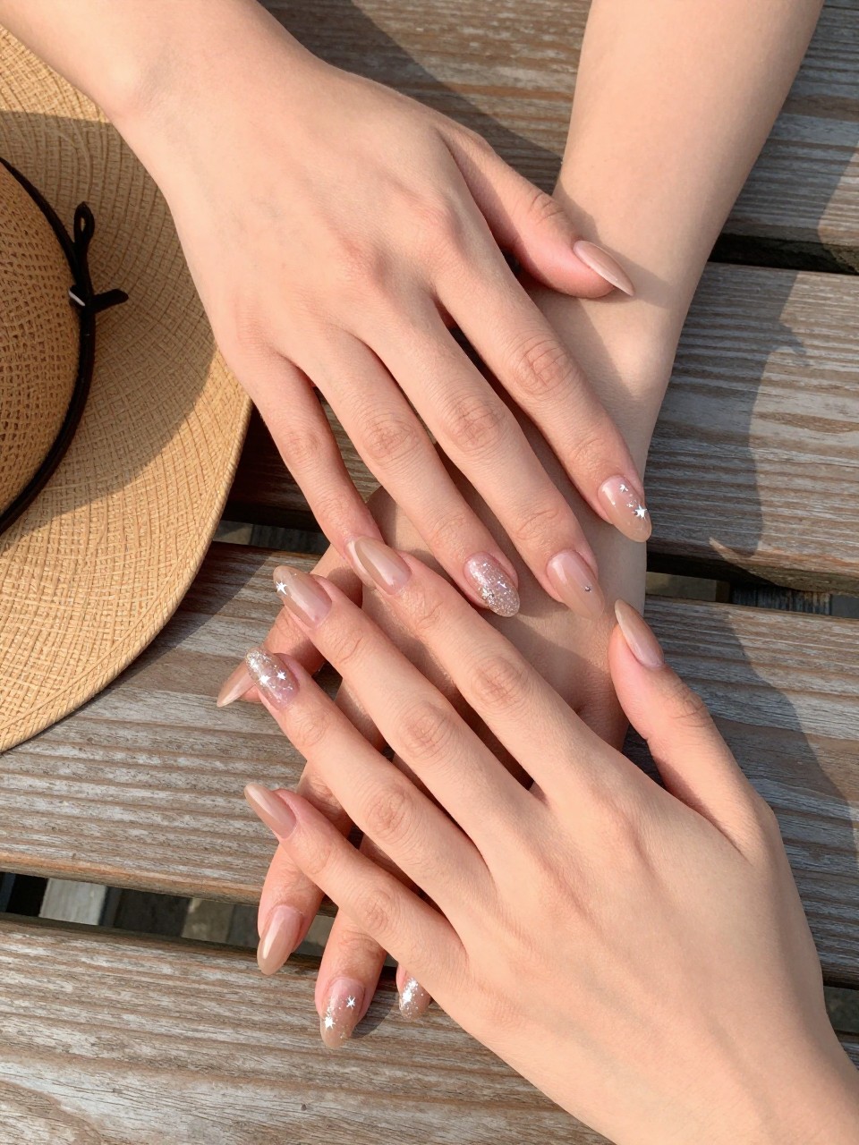 90-degree overhead top-down view of two feminine hands with almond medium nails.
The manicure features a warm nude base with fine glitter overlay and small star accents across two nails.
Include glossy gel finish.

Hands are posed one hand placed gently over the other.

Scene set on a wooden picnic table with a small cowboy hat nearby.

Bright natural daylight with soft shadows, hyper-realistic lifestyle photography, sharp focus on nails, natural skin texture, realistic hands, no extra fingers.