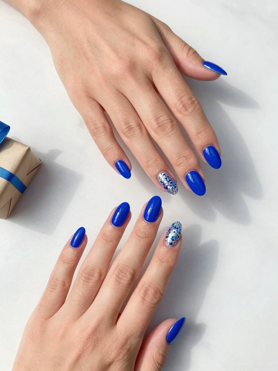 90-degree overhead top-down view of two feminine hands with almond medium nails.
The manicure features a full electric blue base on most nails with one full confetti glitter accent nail on each hand.
Include reflective party glitter with a glossy gel finish.

Hands are posed one hand placed gently over the other.

Scene set on a white marble surface with a small wrapped gift nearby.
Bright natural daylight with soft shadows, hyper-realistic lifestyle photography, sharp focus on nails, natural skin texture, realistic hands, no extra fingers.