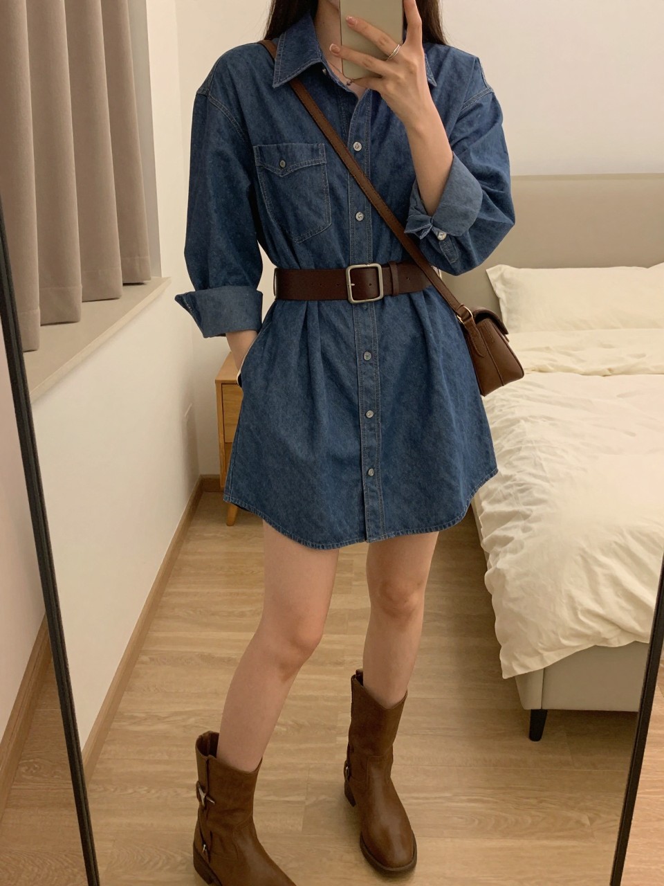 iPhone mirror selfie of a woman wearing a chambray shirt dress cinched with a wide brown leather belt at her waist and matching brown ankle boots, a brown crossbody bag across her body, posed with one hand in her pocket, cropped at NECK so face doesn't show, in a feminine-themed bedroom, warm moody lighting, iPhone photo quality.