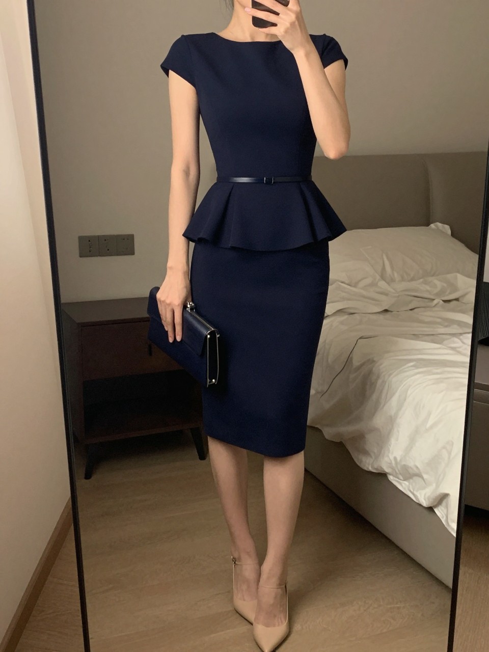 iPhone mirror selfie of a woman wearing a navy peplum dress with a fitted bodice and flared waist detail, nude pointed-toe heels, and a thin belt at her waist, holding a matching clutch, posed with one hand on her hip, cropped at NECK so face doesn't show, in a feminine-themed bedroom, warm moody lighting, iPhone photo quality.