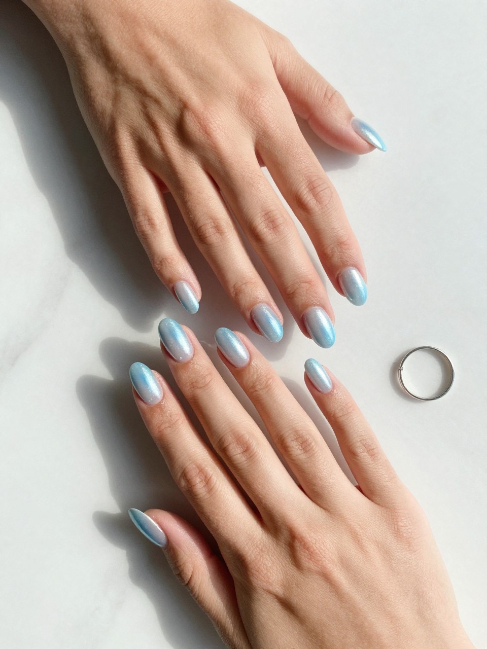 90-degree overhead top-down view of two feminine black hands with almond medium nails.
The manicure features an icy blue base with a glazed chrome overlay creating a pearl-like reflective shine across all nails.
Include glossy gel finish.

Hands are posed one hand placed gently over the other.

Scene set on a white marble surface with a small silver ring nearby.

Bright natural daylight with soft shadows, hyper-realistic lifestyle photography, sharp focus on nails, natural skin texture, realistic hands, no extra fingers.