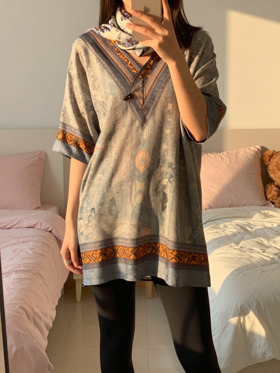 iPhone mirror selfie of a woman in a printed tunic and leggings, holding her phone low while holding a scarf, cropped at NECK so face doesn't show, in a feminine-themed bedroom, evening golden hour, iPhone photo quality.
