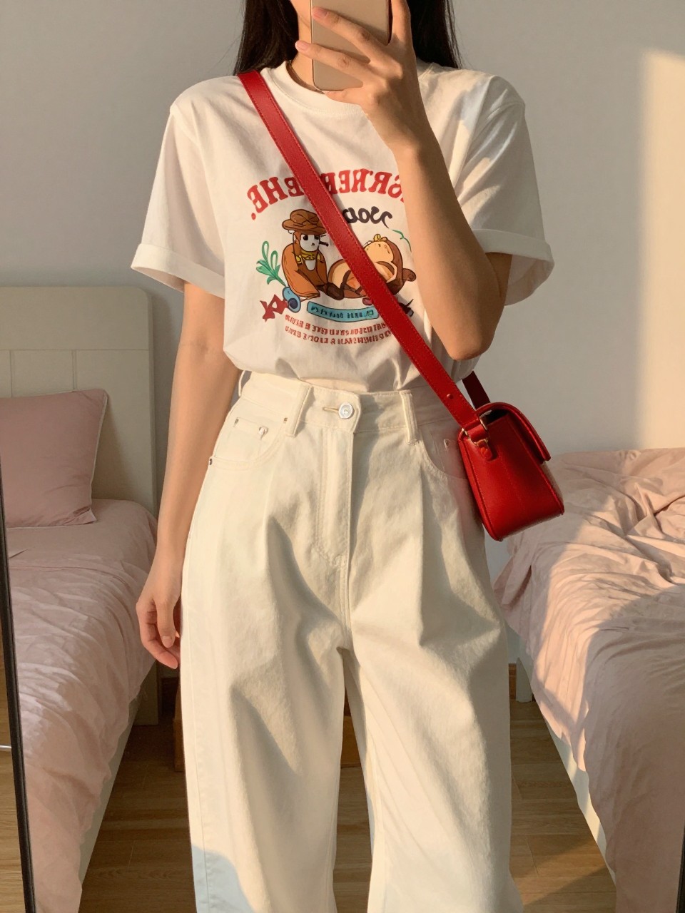 iPhone mirror selfie of a woman in a graphic tee tucked into white wide leg pants with a red crossbody bag, holding phone with slight lean, cropped at NECK so face doesn't show, in a feminine-themed bedroom, evening golden hour, iPhone photo quality.
