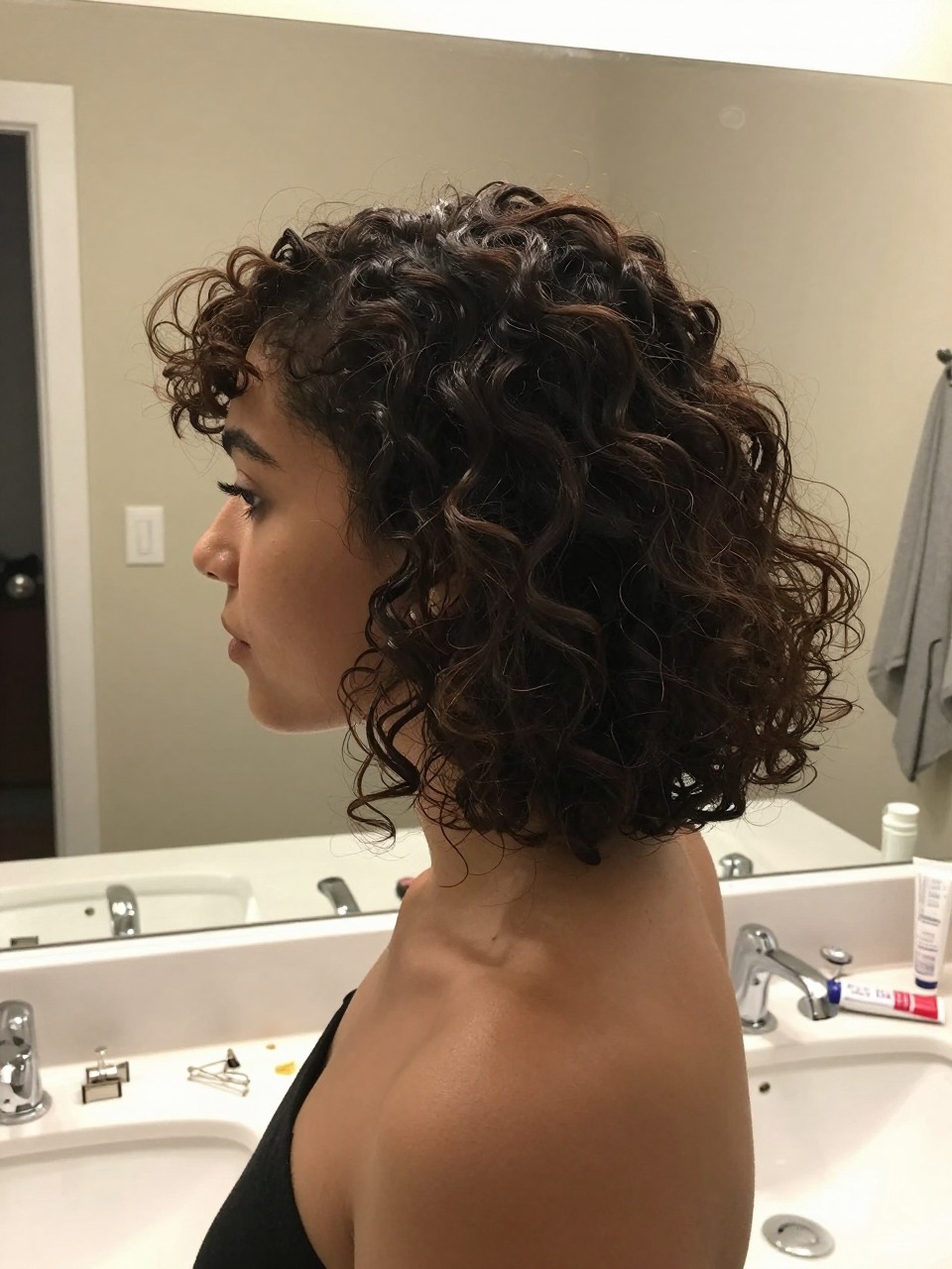 Photo of a woman wearing a low curly bun with soft face-framing curls, side profile view, setting in a bathroom vanity mirror, lighting soft indoor lighting, containing a small lived-in detail such as bobby pins on the counter and a curl cream tube nearby, latest iPhone photo quality.