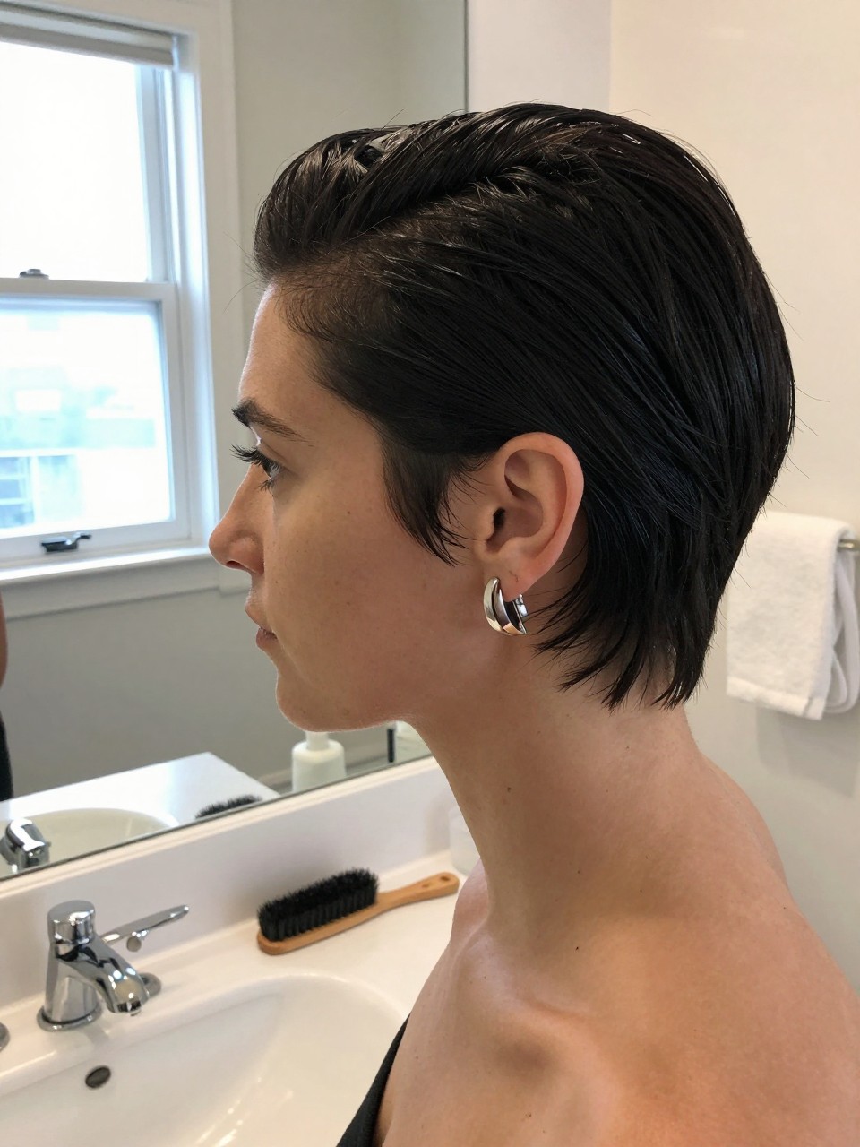 Photo of a woman wearing a slick back pixie with neatly defined sideburn pieces, side profile view, setting in a bathroom vanity mirror, lighting natural window light, containing a small lived-in detail such as an edge brush on the counter and statement earrings peeking through, latest iPhone photo quality.