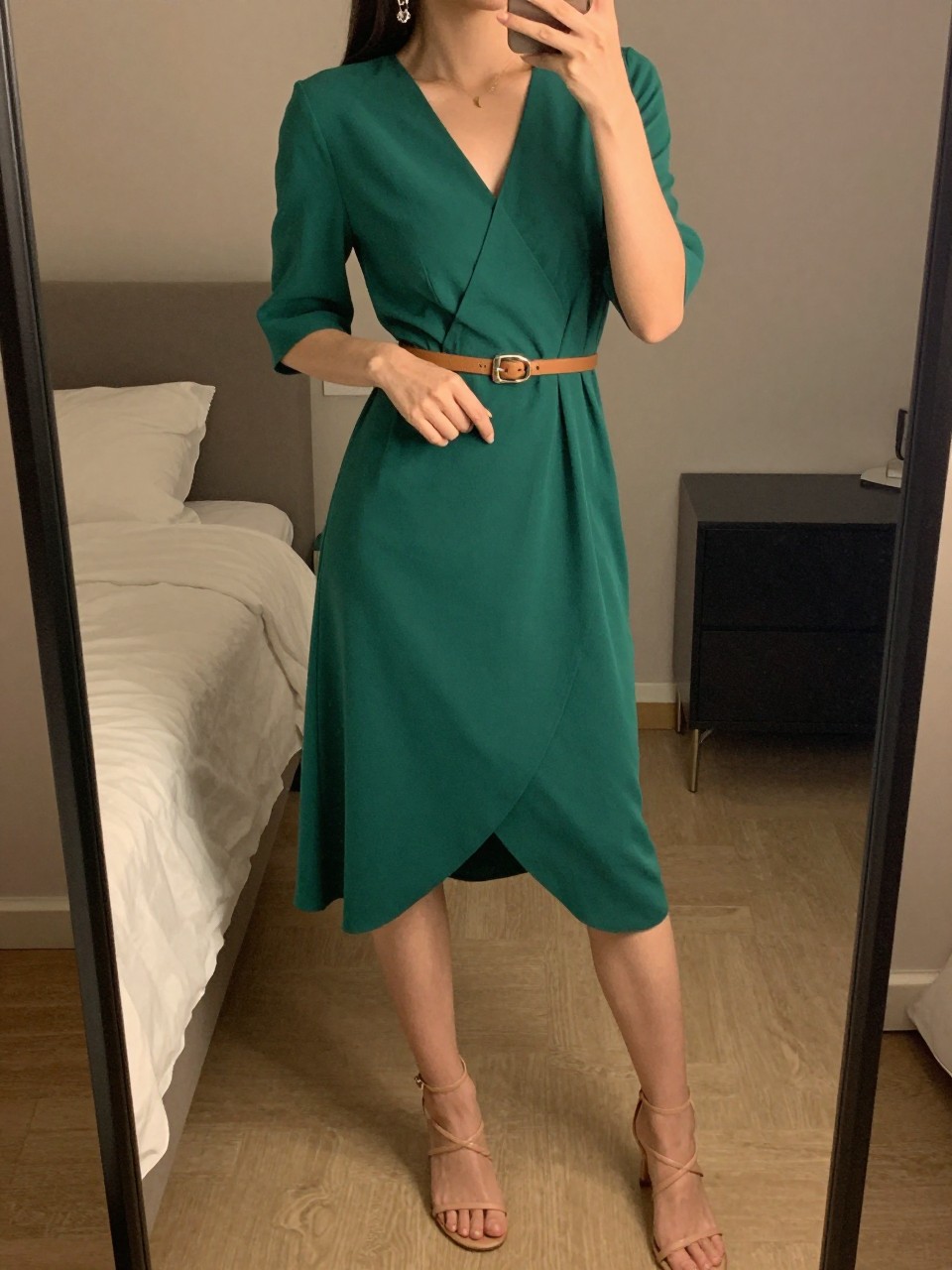 iPhone mirror selfie of a woman wearing a emerald green wrap dress with a thin leather belt cinched at her natural waist and nude strappy heeled sandals, dangling earrings visible, posed with one hand on the belt detail, cropped at NECK so face doesn't show, in a feminine-themed bedroom, warm moody lighting, iPhone photo quality.