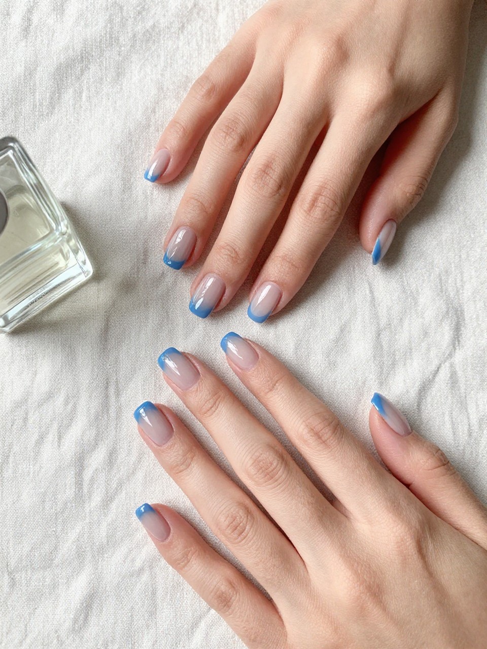 90-degree overhead top-down view of two feminine hands with rounded square medium nails.
The manicure features a sheer blue jelly base with a glossy translucent finish across all nails.
Include smooth gel finish.

Hands are posed fingers gently spread.

Scene set on a soft linen cloth with a small glass perfume bottle nearby.

Bright natural daylight with soft shadows, hyper-realistic lifestyle photography, sharp focus on nails, natural skin texture, realistic hands, no extra fingers.