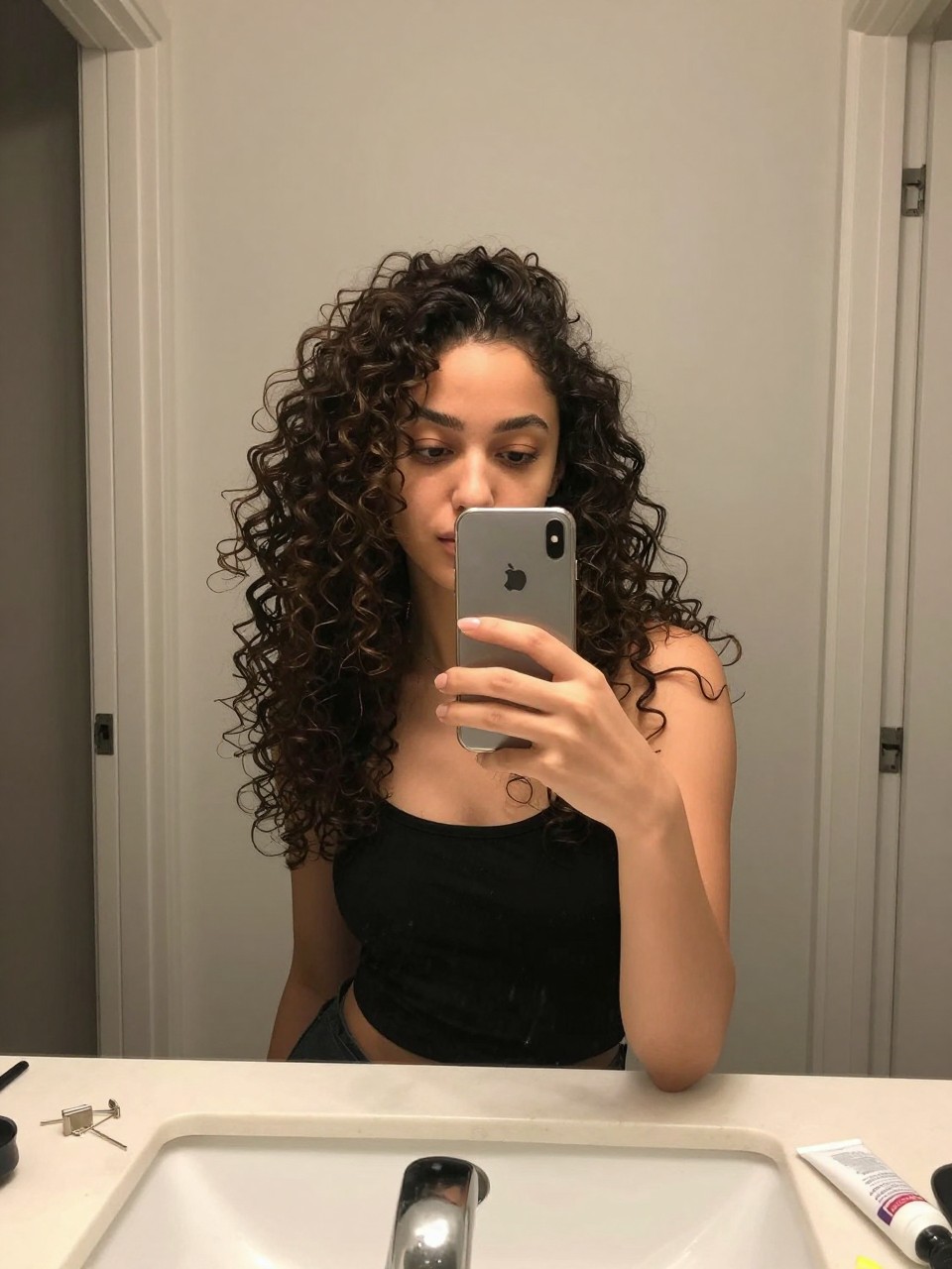 Photo of a woman wearing a curly half-up top knot with defined curls down, candid mirror selfie, setting in a bathroom vanity mirror, lighting soft indoor lighting, containing a small lived-in detail such as bobby pins on the counter and a curl cream tube nearby, latest iPhone photo quality.