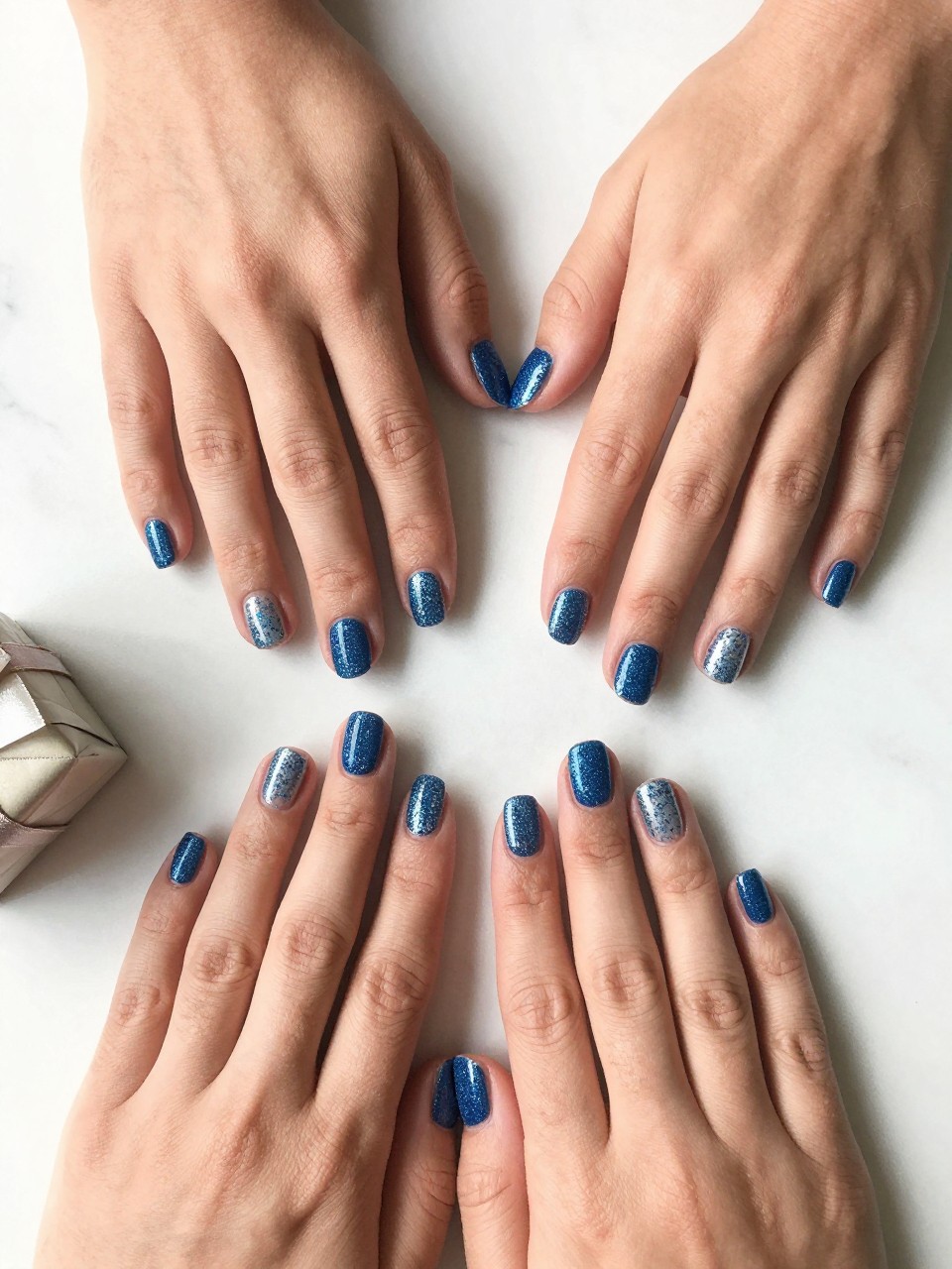 90-degree overhead top-down view of two feminine hands with rounded square medium nails.
The manicure features a blue glitter base on most nails with one full extra-glitter accent nail on each hand.
Include reflective sparkle with a glossy gel finish.

Hands are posed resting flat.

Scene set on a white marble surface with a small wrapped gift nearby.

Bright natural daylight with soft shadows, hyper-realistic lifestyle photography, sharp focus on nails, natural skin texture, realistic hands, no extra fingers.