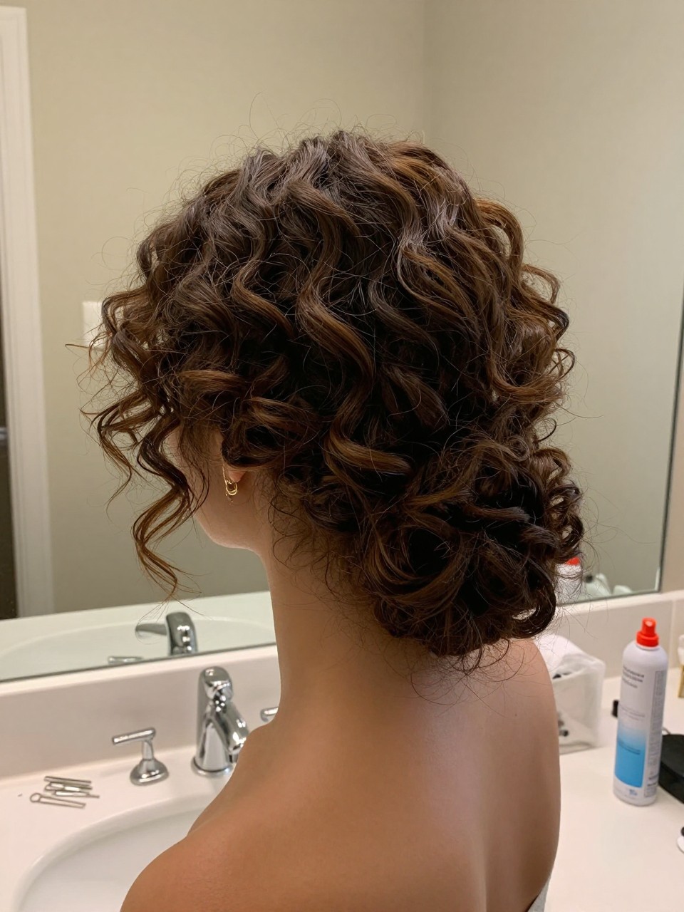 Photo of a woman wearing a curly low chignon with soft volume, back-of-head view, setting in a bathroom vanity mirror, lighting soft indoor lighting, containing a small lived-in detail such as U-pins on the counter and a curl spray bottle nearby, latest iPhone photo quality.