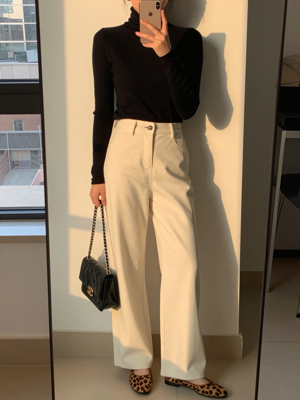 iPhone mirror selfie of a woman in a black turtleneck tucked into soft cream wide leg pants with leopard flats, holding a black chain bag, slight lean against a wall, cropped at NECK so face doesn't show, in a city apartment, evening golden hour, iPhone photo quality.