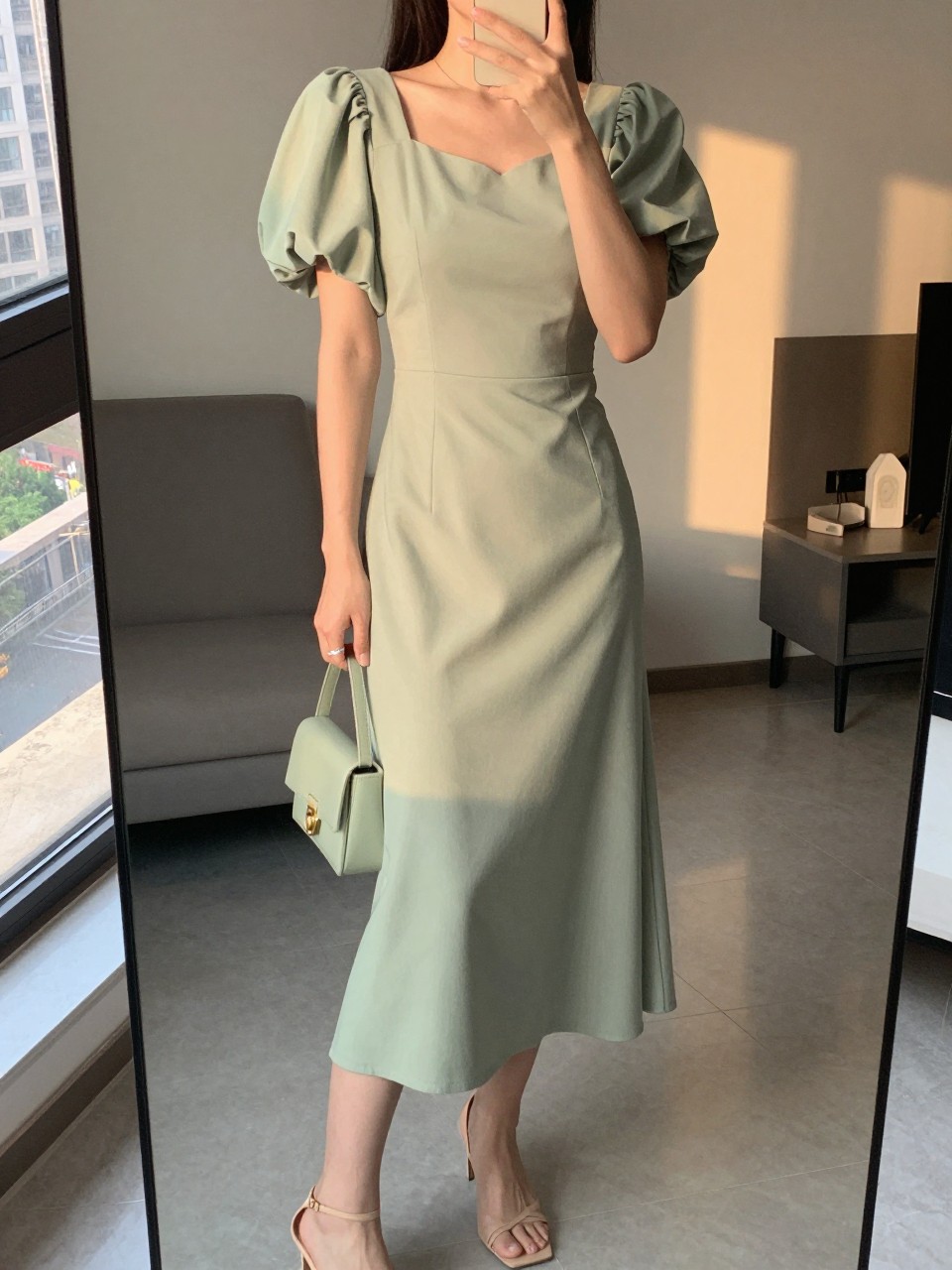 iPhone mirror selfie of a woman wearing a sage green puff sleeve dress with a fitted bodice and nude delicate strappy heels, a mini matching bag, posed with one hand gently touching the puff sleeve, cropped at NECK so face doesn't show, in a city apartment, evening golden hour, iPhone photo quality.