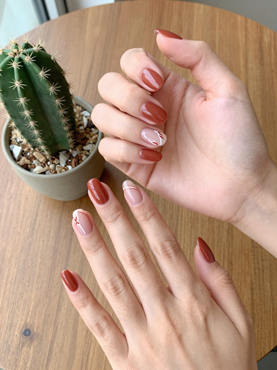 90-degree overhead top-down view of two feminine hands with almond medium nails.
The manicure features a terracotta base with thin white western line art designs on two accent nails.
Include glossy gel finish.

Hands are posed fingers gently spread.

Scene set on a wooden table with a small cactus plant nearby.

Bright natural daylight with soft shadows, hyper-realistic lifestyle photography, sharp focus on nails, natural skin texture, realistic hands, no extra fingers.