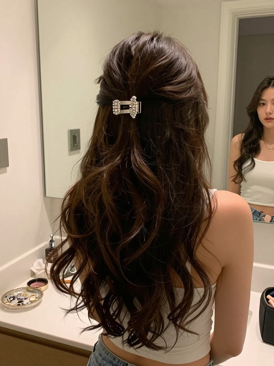 Photo of a woman wearing soft curled long hair half-up secured with a crystal clip, back-of-head view, setting in a bathroom vanity mirror, lighting soft indoor lighting, containing a small lived-in detail such as a jewelry dish on the counter and a makeup bag nearby, latest iPhone photo quality.