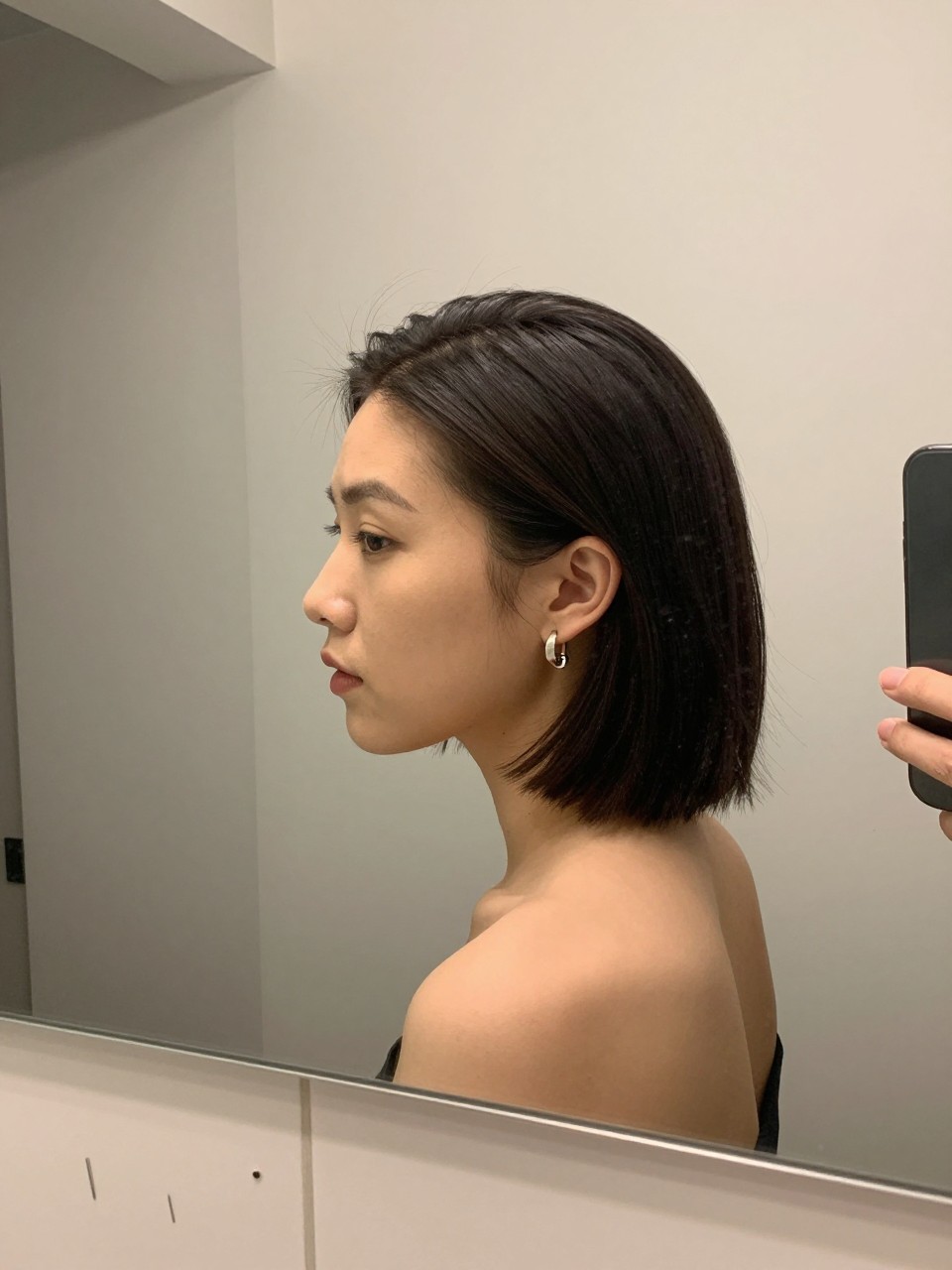 Photo of a woman wearing a sleek middle-part short bob with both sides pinned back behind the ears, side profile view, setting in a bathroom vanity mirror, lighting soft indoor lighting, containing a small lived-in detail such as bobby pins on the counter and statement earrings peeking through, latest iPhone photo quality.