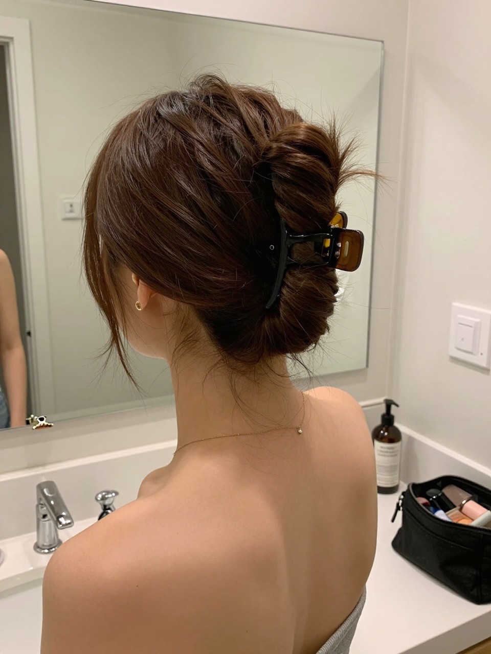 Photo of a woman wearing a claw clip French twist, back-of-head view to show the twist and clip, setting in a bathroom vanity mirror, lighting soft indoor lighting, containing a small lived-in detail such as a claw clip on the counter and a makeup bag nearby, latest iPhone photo quality.
