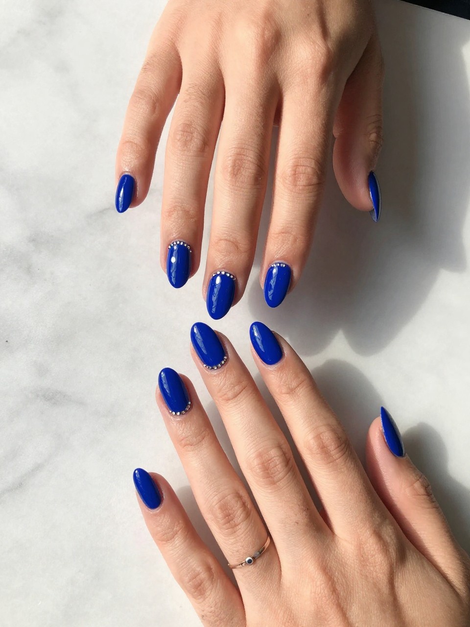 90-degree overhead top-down view of two feminine black hands with almond medium nails.
The manicure features a rich cobalt blue base with a slim line of tiny rhinestones on one accent nail per hand.
Include reflective gemstone details with a glossy gel finish.

Hands are posed fingers gently spread.

Scene set on a marble surface with a small bracelet nearby.

Bright natural daylight with soft shadows, hyper-realistic lifestyle photography, sharp focus on nails, natural skin texture, realistic hands, no extra fingers.