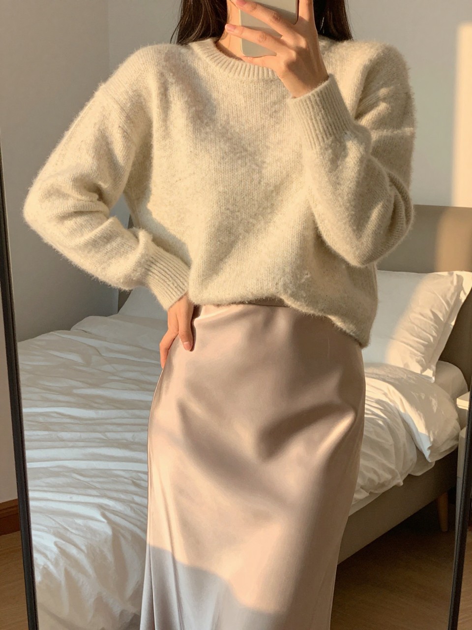 iPhone mirror selfie of a woman in a cozy sweater tucked into a shiny silk skirt, one hand lightly resting on her hip, cropped at NECK so face doesn't show, in a feminine-themed bedroom, evening golden hour, iPhone photo quality.