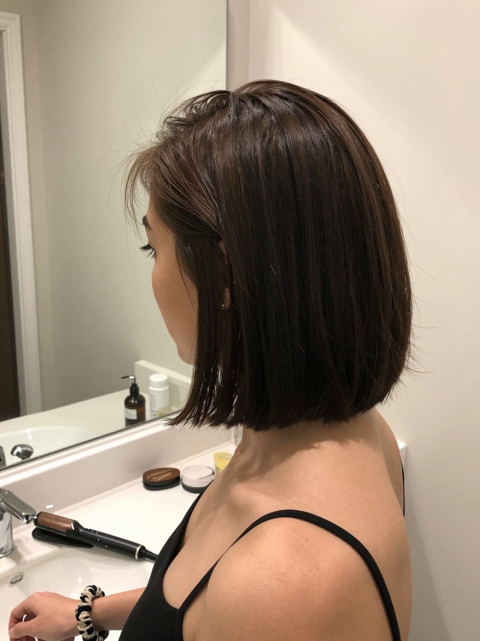 Photo of a woman wearing a slicked back chin-length bob with ends softly curved under, side profile view, setting in a bathroom vanity mirror, lighting soft indoor lighting, containing a small lived-in detail such as a flat iron resting on the counter and a scrunchie on the wrist, latest iPhone photo quality.