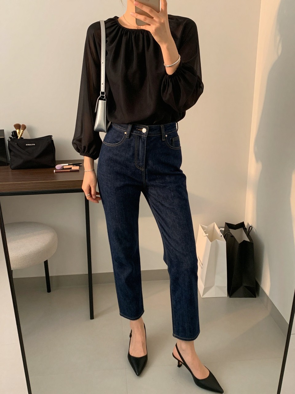 mirror selfie of a woman wearing the exact outfit described above, clearly showing a sheer black floaty blouse, dark indigo tailored denim trousers, black pointed slingback heels, a small silver shoulder bag, and a slim silver bracelet stack, holding a phone in front of a mirror, cropped at NECK so the face does not show, with a relaxed casual selfie pose, in a fashion studio mirror corner, evening golden hour, including makeup brush or cosmetic bag on the counter and shopping bags beside a chair, latest iPhone photo quality. (no watermarks on images)