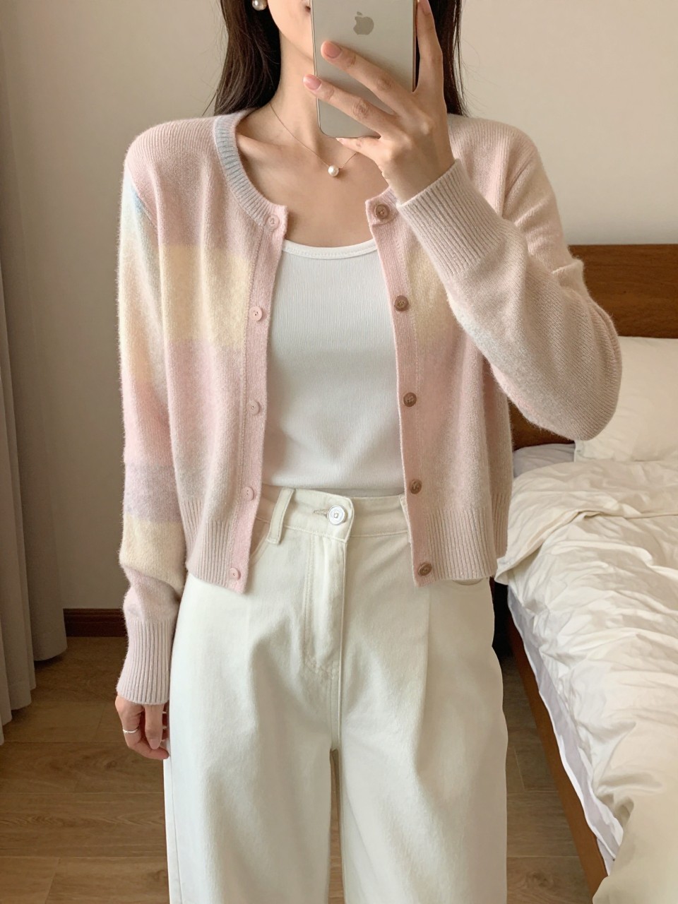 iPhone mirror selfie of a woman in a pastel cardigan over a white tank with white wide leg pants and pearl earrings, hand holding phone at side, cropped at NECK so face doesn't show, in a feminine-themed bedroom, warm moody lighting, iPhone photo quality.