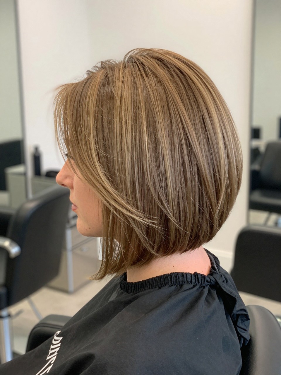 Photo of a white american woman wearing a cropped A-line bob with a smooth outline, stacked back, and longer front corners, with the haircut fully visible and clearly defined, shot from a salon mirror reveal angle, in a bright salon interior, with soft indoor lighting, showing the haircut shape, layers, ends, bangs, and texture clearly, including the salon chair edge, latest iPhone photo quality.