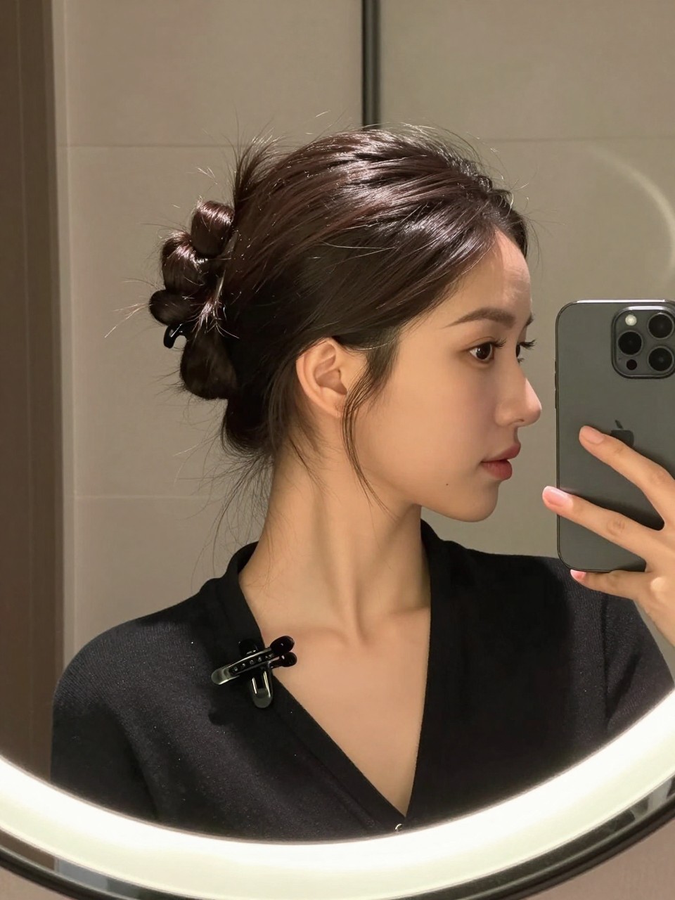 Photo of a woman wearing a slicked back top knot, side profile mirror selfie showing the rounded bun at the crown, setting is a bathroom vanity mirror with soft indoor lighting, containing a hair clip clipped to her collar, iPhone photo quality.